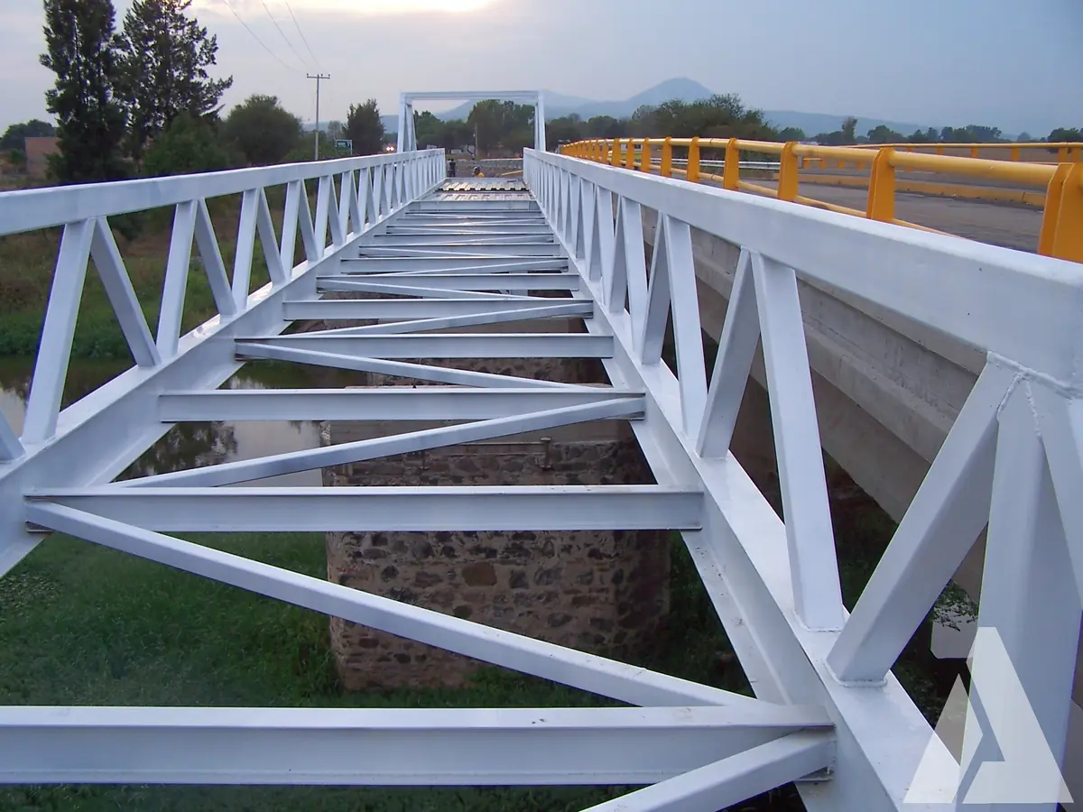 Armadura metálica triangulada pintada de blanco de un puente peatonal apoyada en pilas de piedra sobre un cauce